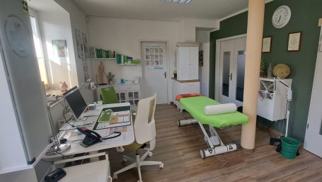 Praxisraum mit Schreibtisch und Bürostuhl, hellgrün bezogener Behandlungsliege, diverse weiße Praxis-Möbel im Hintergrund