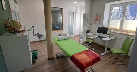 Praxisraum mit mittiger, hellgrün bezogener Behandlungslieg, Schreibtisch und hellgrünem Besucherstuhl. Fenster mit Blick in den Hof