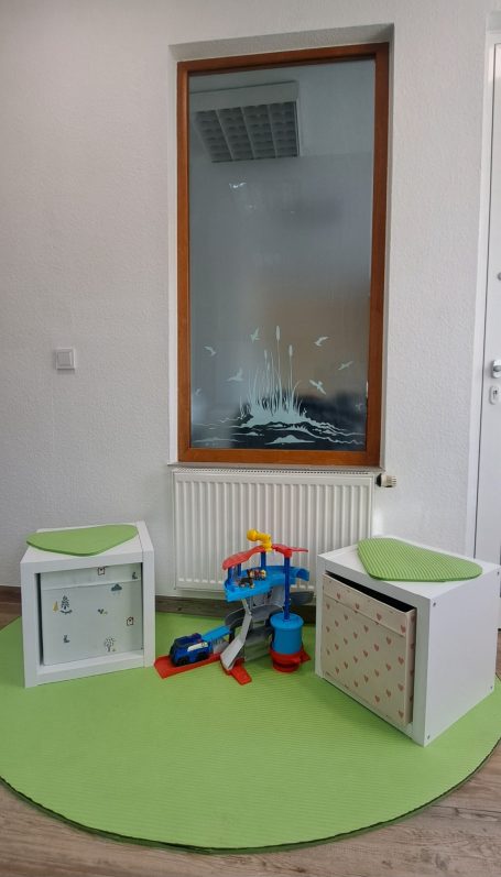 Zwei kleine quadratische Sitzhocker und Spielgeräte auf einer hellgrünen Unterlage vor einem Innenfenster mit transparentem Strandmotiv
