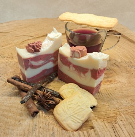 Zwei Seifenstücke auf einer grob geschnittenen Holzscheibe, dekoriert mit weihnachtlichen Gewürzen, Spekulatius und einem Glas mit Glühwein